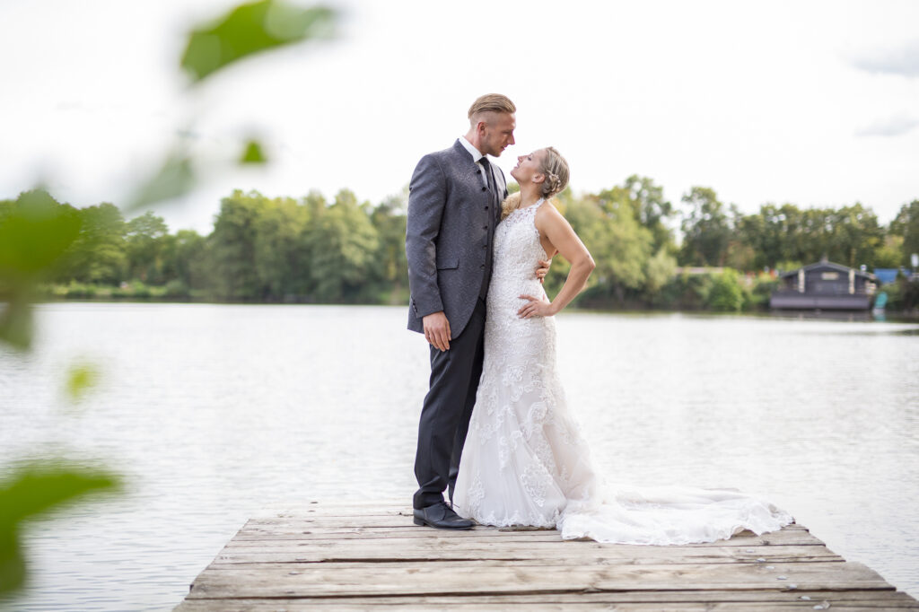 Brautpaar steht auf einem Steg eines Sees, im Hintergrund erkannt man unscharf ein Bootshaus sowie den See und dessen Ufer