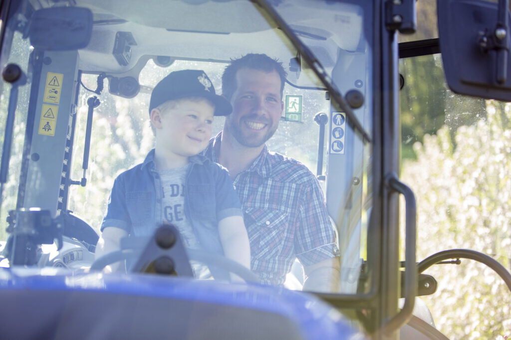 Vater und Sohn sitzen auf einem Traktor, der Sohn sitzt auf dem Schoß des Vaters, Blick durch die Windschutzscheibe