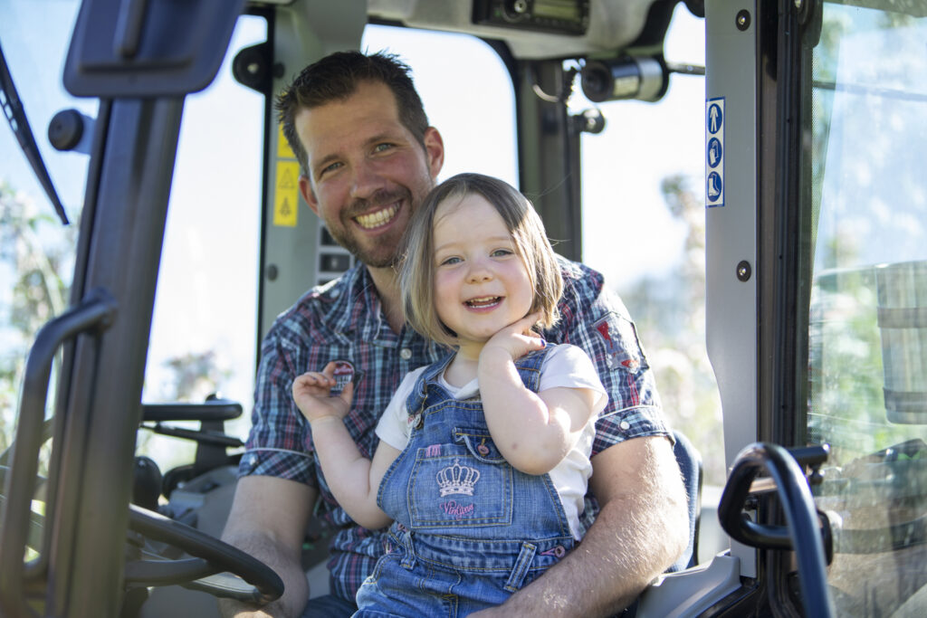 Vater und Tochter sitzen auf einem Traktor, beide schauen in die Kamera, die Tochter sitzt auf dem Schoß des Vaters