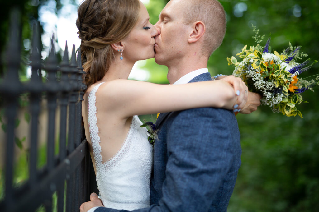 Brautpaar küsst sich, sie ist an einen Metallzaun gelehnt und hält ihren Strauß in der Hand