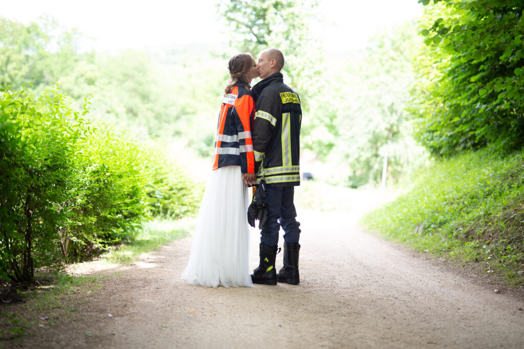 Hochzeitspaar küsst sich, er trägt eine Feuerwehr-Jacke, sie eine Jacke einer Rettungssanitäterin, sie stehen im Grünen